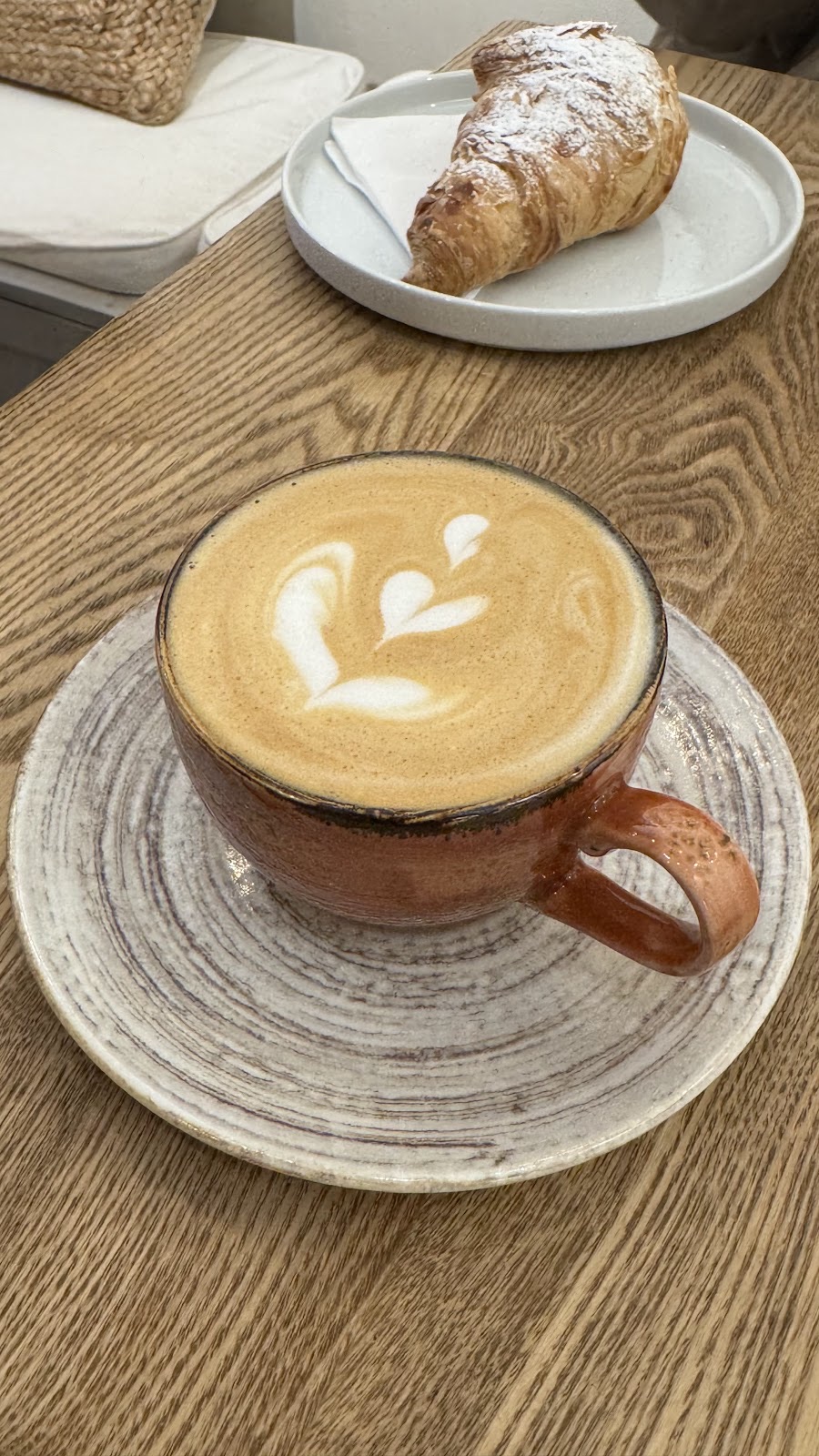A latte in a terracotta cup with a croissant on a plate behind it